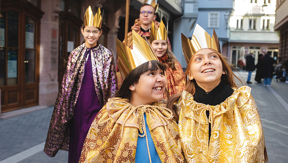 Foto von 4 Sternsingern und einer Begleitperson, alle Kinder tragen eine goldene Krone und einen prachtvollen Umhang. Die Kinder grinsen und lachen.